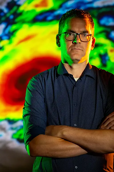 A serious man in a dark blue shirt and glasses stands with his arms crossed in front of a colorful hurricane projection.